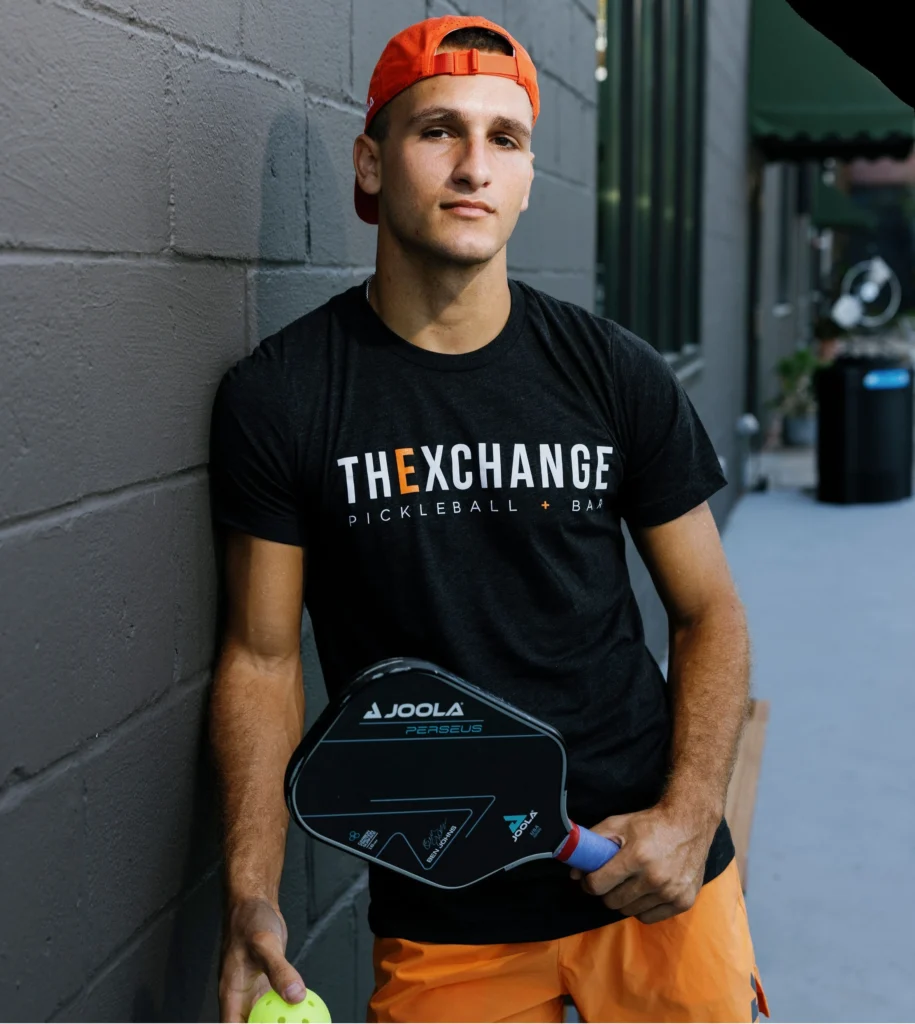 AJ Parfait wearing a The Exchange Pickleball and Bar shirt while holding a pickleball paddle
