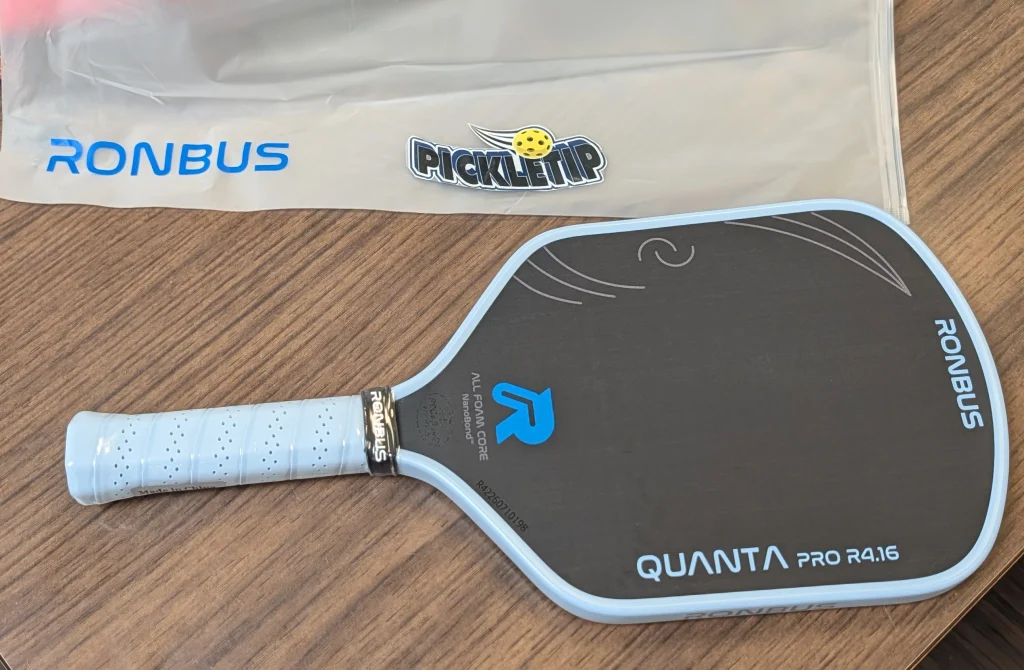 Ronbus Quanta Pro R4.16 paddle on a wooden table beneath a Ronbus sleeve with a PickleTip sticker.